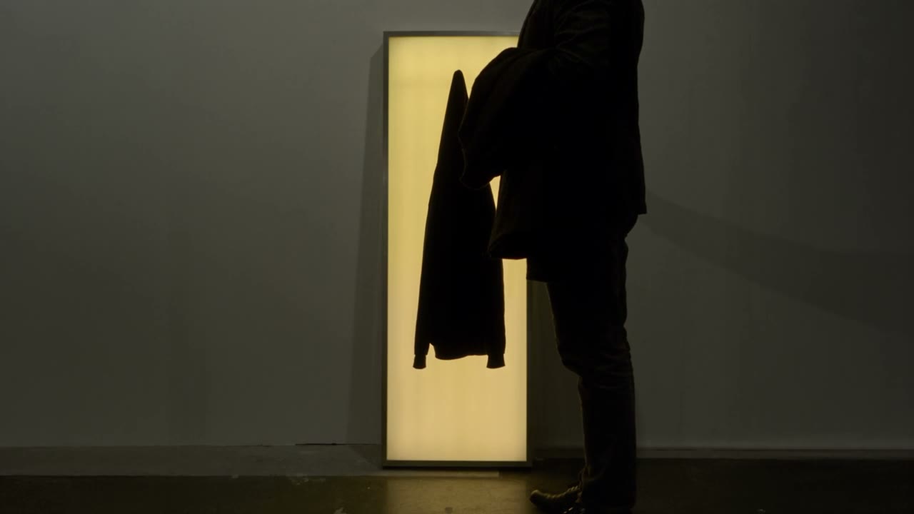 Warm Welcome: a tall light-box coat stand by a wall; a coat hung on it becomes a dark silhouette against warm light.